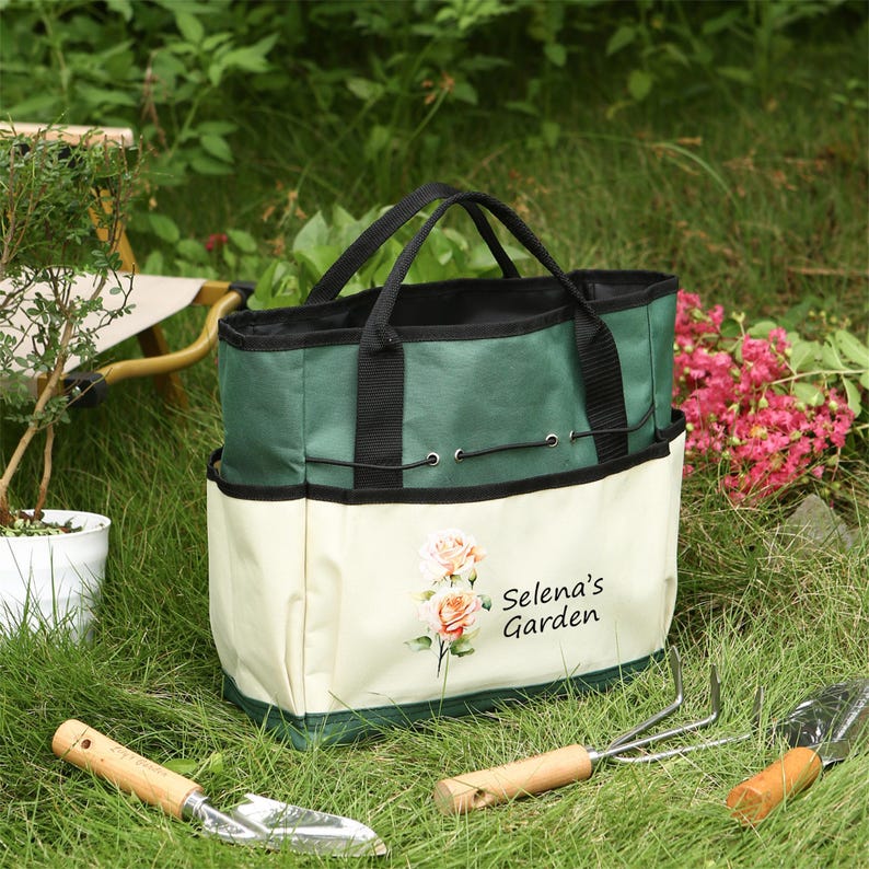 Puede incluir: Bolsa de jard&iacute;n de lona verde y crema con asas negras y m&uacute;ltiples bolsillos. La bolsa tiene un dise&ntilde;o floral y el texto "Selena's Garden" impreso en la parte delantera.