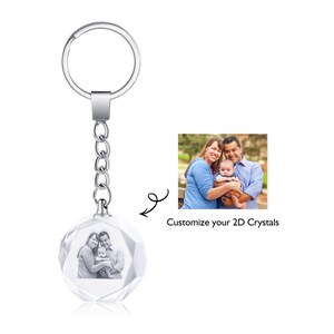 May include: A silver keychain with a crystal charm that can be customized with a 2D photo. The charm features a family of three, a couple holding a baby. The text "Customize your 2D Crystals" is visible below the image.