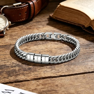 Könnte beinhalten: Ein silbernes Edelstahlarmband mit einem Panzerketten-Design. Das Armband verfügt über drei rechteckige Namensschilder mit den eingravierten Namen "Selena", "Daniel" und "Matthew". Das Armband wird auf einer Holzoberfläche präsentiert.