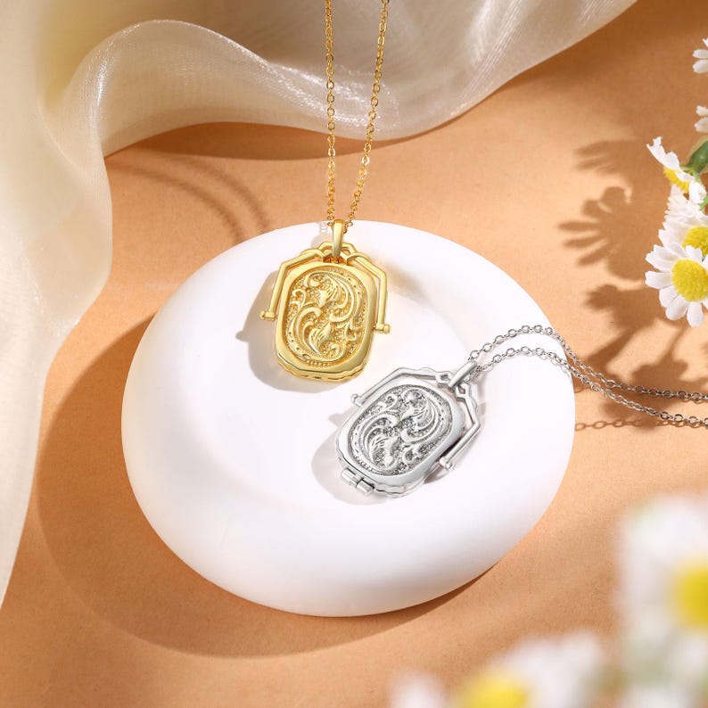 May include: Two ornate pendant necklaces. One is gold-toned, the other silver-toned. Both feature a rectangular design with intricate floral patterns. The pendants hang from delicate chains, resting on a white, circular surface.