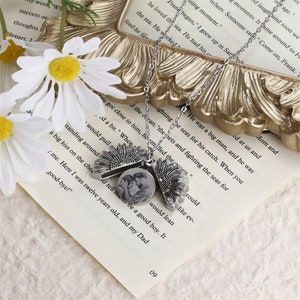 Pu&ograve; includere: Una collana color argento con un ciondolo a forma di girasole che si apre per rivelare una piccola foto. La collana include anche due ciondoli a forma di girasole. Poggia su una pagina di libro con testo.