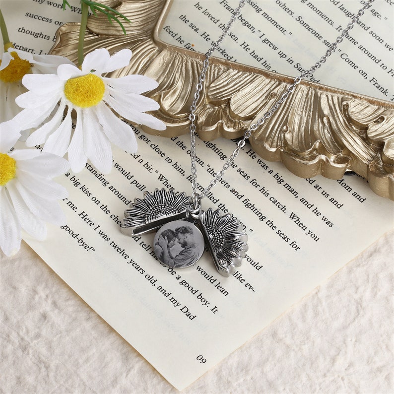 Pu&ograve; includere: Una collana color argento con un ciondolo a forma di girasole che si apre per rivelare una piccola foto. La collana include anche due ciondoli a forma di girasole. Poggia su una pagina di libro con testo.