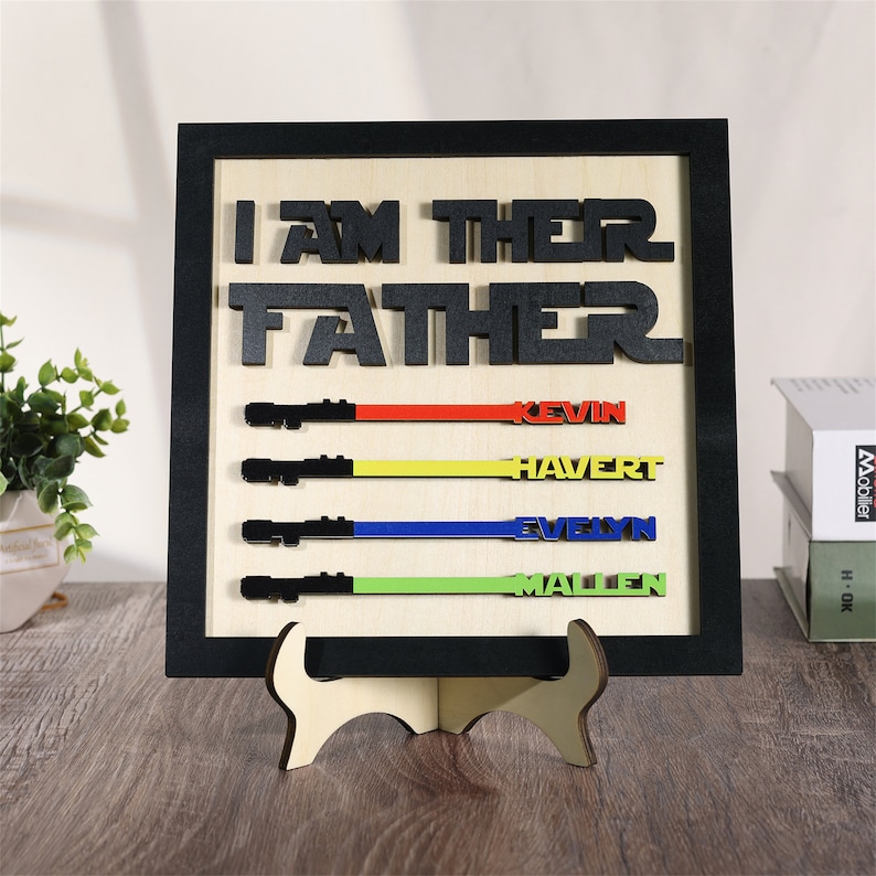 Puede incluir: Un letrero de madera con el texto "I AM THE FATHER" en letras negras. El letrero tiene cuatro piezas en forma de sable de luz con rayas de diferentes colores y nombres: Kevin, Havert, Evelyn y Mallen.