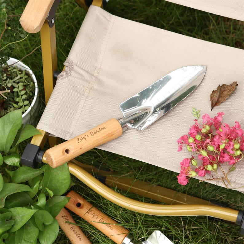 Puede incluir: Un primer plano de una azada de jard&iacute;n con mango de madera. El mango est&aacute; grabado con las palabras "Lily's Garden". La azada est&aacute; apoyada en una silla de lona beige.