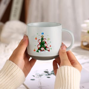 Peut inclure: Une tasse en céramique blanche avec un motif d'arbre de Noël. L'arbre est décoré de petites décorations et porte les noms des membres de la famille. La tasse a une poignée et indique "Joyeux Noël" au bas.