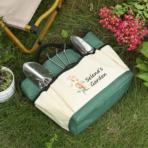Puede incluir: Una bolsa de jard&iacute;n verde y beige con una parte superior con cremallera y varios bolsillos. La bolsa tiene un dise&ntilde;o floral y el texto "Selena's Garden" impreso en la parte delantera.