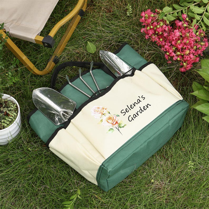 Puede incluir: Una bolsa de jard&iacute;n verde y beige con una parte superior con cremallera y varios bolsillos. La bolsa tiene un dise&ntilde;o floral y el texto "Selena's Garden" impreso en la parte delantera.