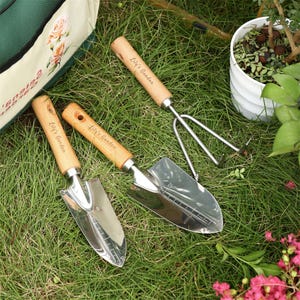 Puede incluir: Tres herramientas de jardiner&iacute;a con mangos de madera y cabezales de metal. Los mangos est&aacute;n grabados con las palabras "Lily's Garden". Las herramientas son una pala, un rastrillo de mano y una pala peque&ntilde;a.