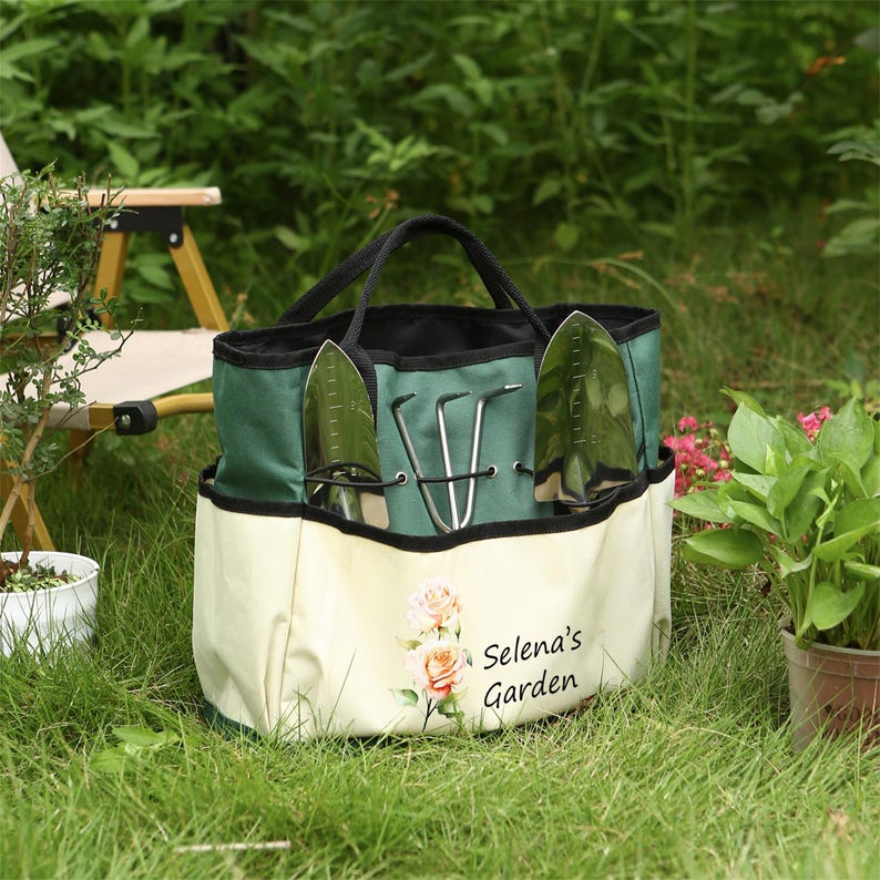 Puede incluir: Una bolsa de jard&iacute;n de lona verde y blanca con m&uacute;ltiples bolsillos. La bolsa tiene un asa negra y est&aacute; decorada con un dise&ntilde;o floral y el texto "Selena's Garden".