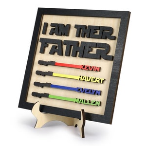 Puede incluir: Un letrero de madera con el texto "Soy su padre" en letras negras. El letrero tiene cuatro sables de luz con los nombres Kevin, Havert, Evelyn y Mallen escritos en ellos en diferentes colores.