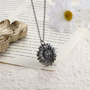 Pu&ograve; includere: Una collana con pendente a forma di girasole color argento. Il pendente presenta un design a girasole dettagliato e tridimensionale. La catena della collana &egrave; anch'essa color argento. La collana poggia su una pagina di libro con una margherita sullo sfondo.