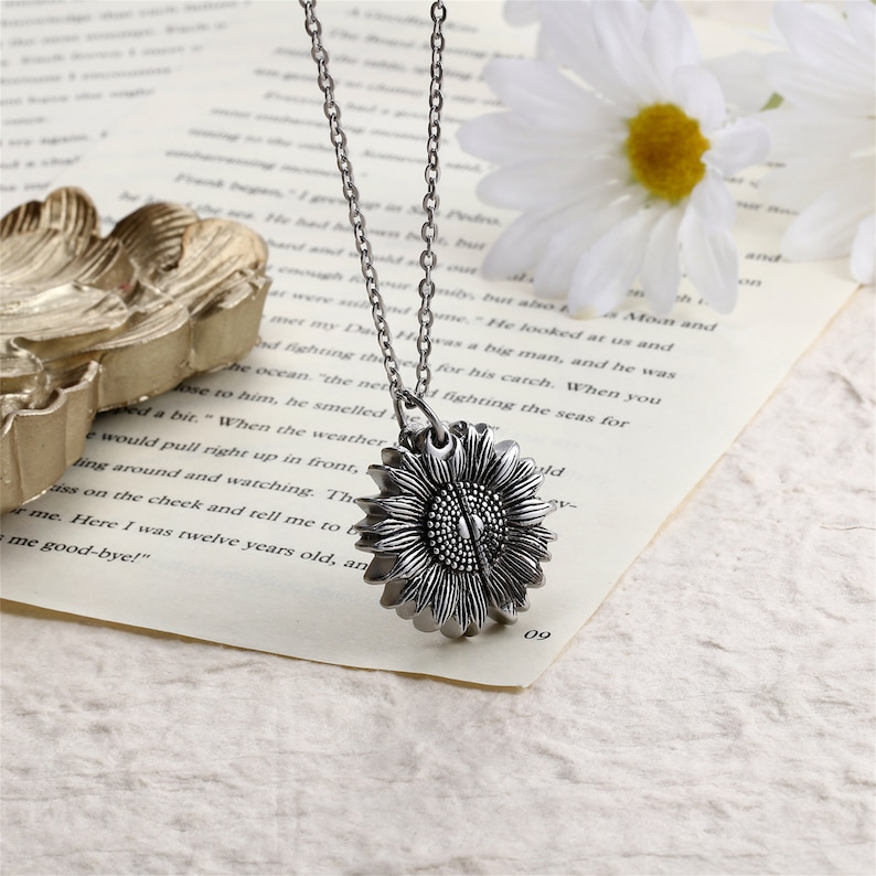 Pu&ograve; includere: Una collana con pendente a forma di girasole color argento. Il pendente presenta un design a girasole dettagliato e tridimensionale. La catena della collana &egrave; anch'essa color argento. La collana poggia su una pagina di libro con una margherita sullo sfondo.