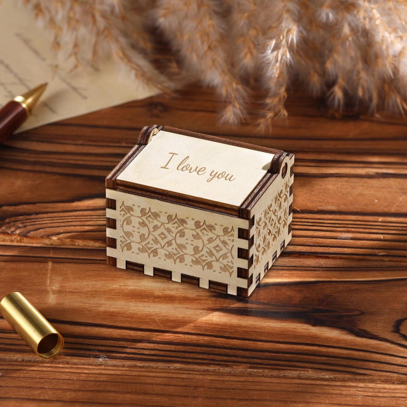Pu&ograve; includere: Un piccolo carillon rettangolare in legno con le parole "I love you" incise sul coperchio. La scatola presenta intricati motivi floreali e un bordo marrone scuro. La scatola &egrave; appoggiata su una superficie di legno.