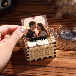 Pu&ograve; includere: Un piccolo carillon in legno con una foto di una coppia innamorata. La scatola &egrave; rettangolare con un motivo floreale decorativo e un meccanismo a forma di tastiera. Note musicali illustrate sopra la scatola.
