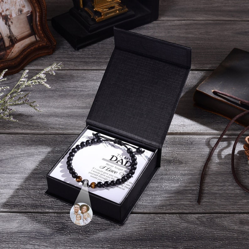 Pu&ograve; includere: Una scatola regalo nera con un braccialetto nero a perline all'interno. Il braccialetto ha un piccolo ciondolo d'argento con una foto di famiglia. La scatola contiene un biglietto bianco con il testo "To My Dad Never forget that I love you" su di esso.