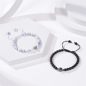 Pu&ograve; includere: Due braccialetti con perline, uno bianco con accenti neri e uno nero con accenti argentati. Entrambi i braccialetti hanno un ciondolo argentato al centro.