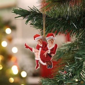 Peut inclure: Une décoration de Noël représentant un couple habillé en Père Noël. La femme est assise sur les genoux de l'homme et ils sourient tous les deux. La décoration est en acrylique transparent et possède un cordon doré pour l'accrocher.