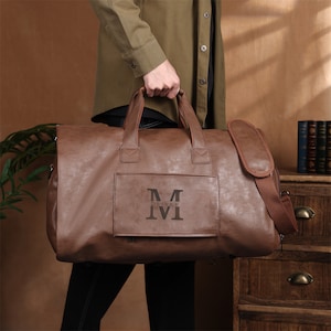 May include: A brown leather duffel bag with a shoulder strap and top handles. The bag features a rectangular pocket with the name "OLIVER" and the initial "M" printed on it. The bag is being held by a person.