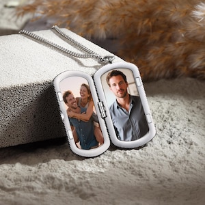 May include: A silver locket necklace with a rectangular pendant that opens to reveal two photographs. One photo shows a couple, and the other shows a man. The locket is on a textured, light-coloured surface, with a silver chain.