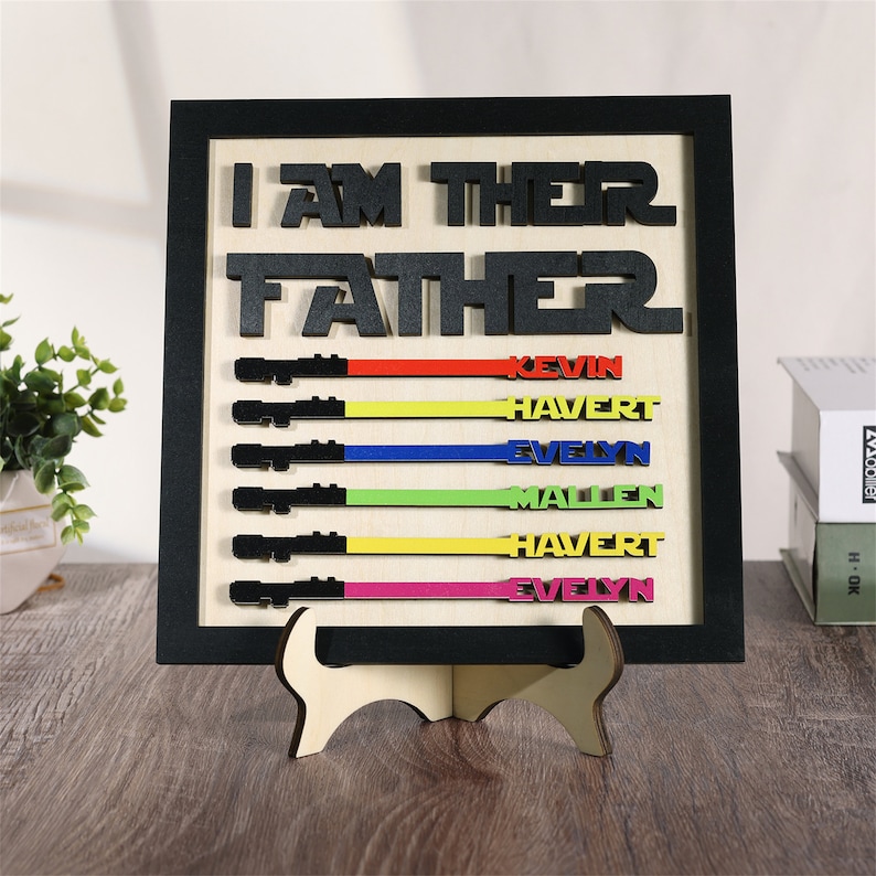 Puede incluir: Un letrero de madera enmarcado en negro con el texto "Soy su padre" en letras negras. El letrero tiene seis sables de luz con rayas de diferentes colores y nombres debajo de cada uno: Kevin, Havert, Evelyn, Mallen, Havert, Evelyn.