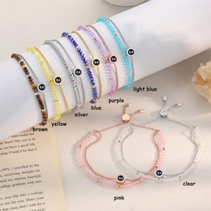 May include: A set of ten bracelets with different coloured beads and a black charm. The bracelets are arranged in a row from left to right, with the colours brown, yellow, silver, blue, purple, light blue, clear, and pink. The bracelets are all adjustable and have a sliding knot.