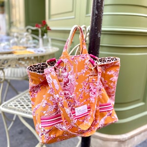 May include: Orange tote bag with a pink floral design and a leopard print lining. The bag has pink handles and a shoulder strap. The bag has a label that says "Madeleine".
