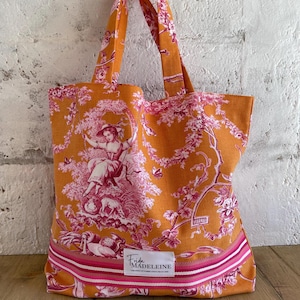 May include: Orange tote bag with a pink toile pattern depicting a pastoral scene. The bag has matching handles and a pink and white striped border. The label reads "Frida Madeleine".