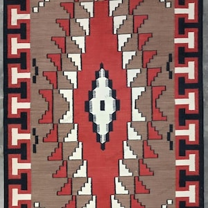 Op de afbeelding: Een Native American tapijt met een geometrisch patroon in rood, wit, zwart en bruin. Het tapijt heeft een rand met een herhalend patroon.