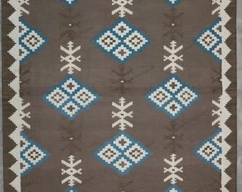 May include: A rectangular area rug featuring a dark brown background with a repeating pattern of blue and white geometric diamond shapes and snowflake-like motifs. The rug is finished with a white zigzag border and white fringe on both ends.