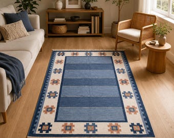 May include: A rectangular area rug featuring a central panel of alternating horizontal blue stripes. The border is cream-colored with a repeating pattern of small, geometric, rust-colored and blue cross-shaped motifs, finished with fringe on both ends.
