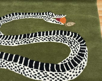 Puede incluir: Una alfombra verde con un diseño de serpiente blanca y negra. La serpiente está enroscada en un círculo y tiene una manzana roja en la boca.