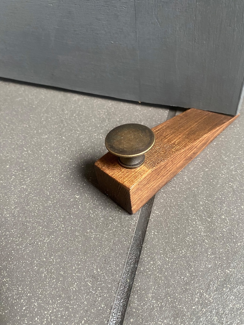 May include: A wooden doorstop with a brass knob. The doorstop is brown and has a natural wood finish. The knob is round and has a shiny finish.