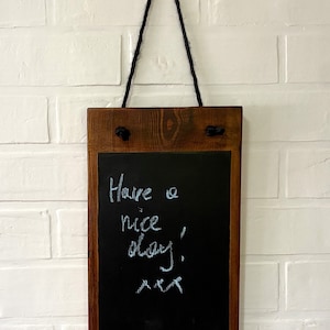 May include: A wooden framed chalkboard with a black surface and a handwritten message that says "Have a nice day!" hanging on a white brick wall with a black rope.