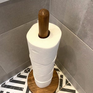 K&ouml;nnte beinhalten: Ein freistehender Toilettenpapierhalter aus Holz mit brauner Oberfl&auml;che. Der Halter hat einen runden Sockel und einen zylindrischen Pfosten. Eine Rolle wei&szlig;es Toilettenpapier befindet sich auf dem Pfosten.