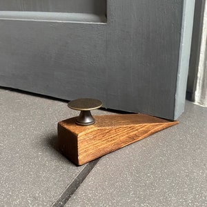 May include: A wooden doorstop with a brass knob. The doorstop is brown and has a wedge shape. It is sitting on a gray tiled floor.