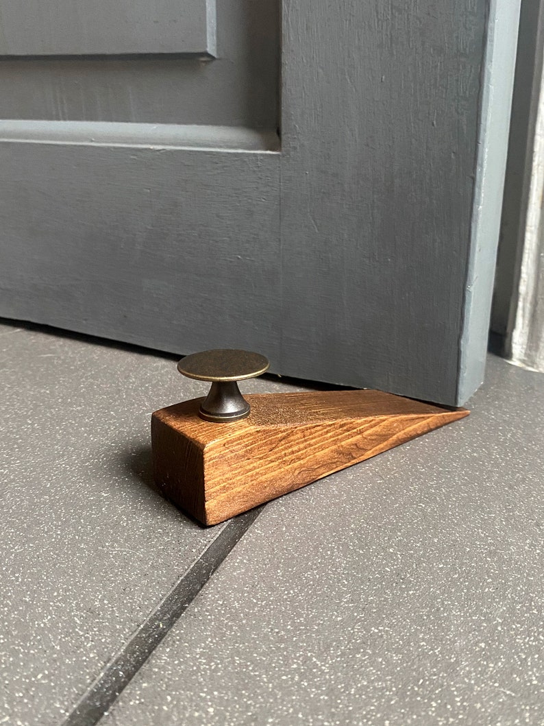 May include: A wooden doorstop with a brass knob. The doorstop is brown and has a wedge shape. It is sitting on a gray tiled floor.