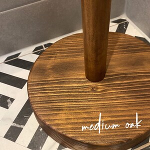 K&ouml;nnte beinhalten: Ein Papierrollenhalter aus Holz mit einem runden Sockel. Der Sockel ist in einem mittleren Eichenholzton gebeizt und tr&auml;gt die Aufschrift "medium oak" in wei&szlig;er Farbe.