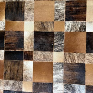 May include: A close-up of a patchwork rug with a geometric pattern. The rug features squares of varying shades of brown, black, and cream, with visible hair texture. The design creates a textured, rustic aesthetic.
