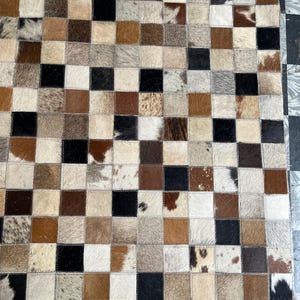 Puede incluir: Una alfombra de patchwork con un patrón de tablero de ajedrez. Los cuadrados están hechos de varios tonos de piel de animal marrón, negro y blanco. La alfombra tiene una estética natural y rústica, adecuada para una sala de estar o dormitorio.