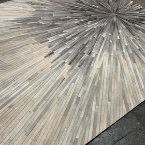 May include: A grey and white cowhide rug with a geometric pattern. The rug is made of strips of leather that are arranged in a radial pattern, creating a sunburst effect.