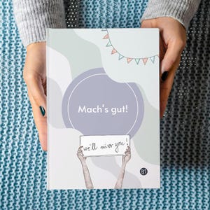 Könnte beinhalten: Eine weiße Karte mit dem Text "Mach's gut!" und "we'll miss you!" darauf. Die Karte wird von zwei Händen gehalten, wobei der Text zum Betrachter zeigt. Die Karte ist mit einem lila Kreis und einer Girlande aus bunten Flaggen verziert.