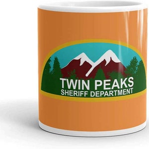 Op de afbeelding: Oranje keramische mok met een witte binnenkant. De mok heeft een groen en blauw ovaal ontwerp met een berglandschap en de woorden "Twin Peaks Sheriff Department" in het wit.