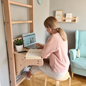 May include: A woman sits at a wooden desk with a laptop, book, and phone. The desk is attached to a wooden wall-mounted pull-up bar. The room has light wood floors and a blue armchair.