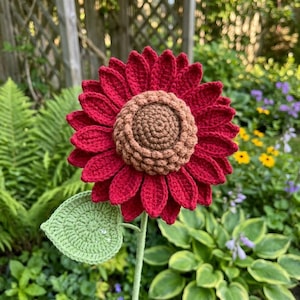 May include: A handcrafted crochet sunflower with deep red petals and a brown center. The flower has a green stem and a green leaf. The sunflower is displayed outdoors in a garden setting.