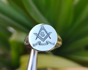 May include: A silver ring with a Masonic symbol engraved on the face. The symbol is a square and compass with a 'G' in the center. The ring also has a banner with the words 'Faith, Hope, Charity' inscribed on it.