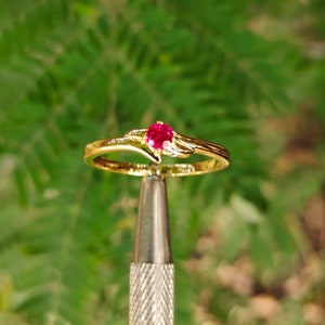 May include: A gold ring with a single red gemstone set in the center. The ring has a delicate, curved design.
