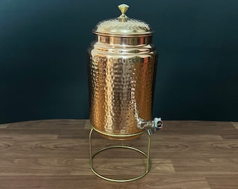 Könnte beinhalten: Ein gehämmerter Kupfer-Wasserdispenser mit einem goldfarbenen Deckel und einem Hahn. Der Dispenser steht auf einem goldfarbenen Metallständer.