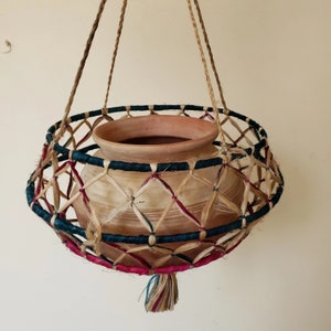 May include: A terracotta pot with a woven hanging basket. The basket is made of natural fibers and has a blue, pink, and brown color scheme. The pot is suspended from a rope.