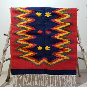K&ouml;nnte beinhalten: Ein roter, blauer, gelber und grauer geometrisch gemusterter Teppich mit Fransen. Der Teppich hat ein traditionelles Design mit einer zentralen Diamantform.