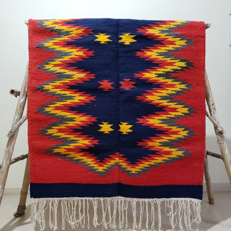 K&ouml;nnte beinhalten: Ein roter, blauer, gelber und grauer geometrisch gemusterter Teppich mit Fransen. Der Teppich hat ein traditionelles Design mit einer zentralen Diamantform.